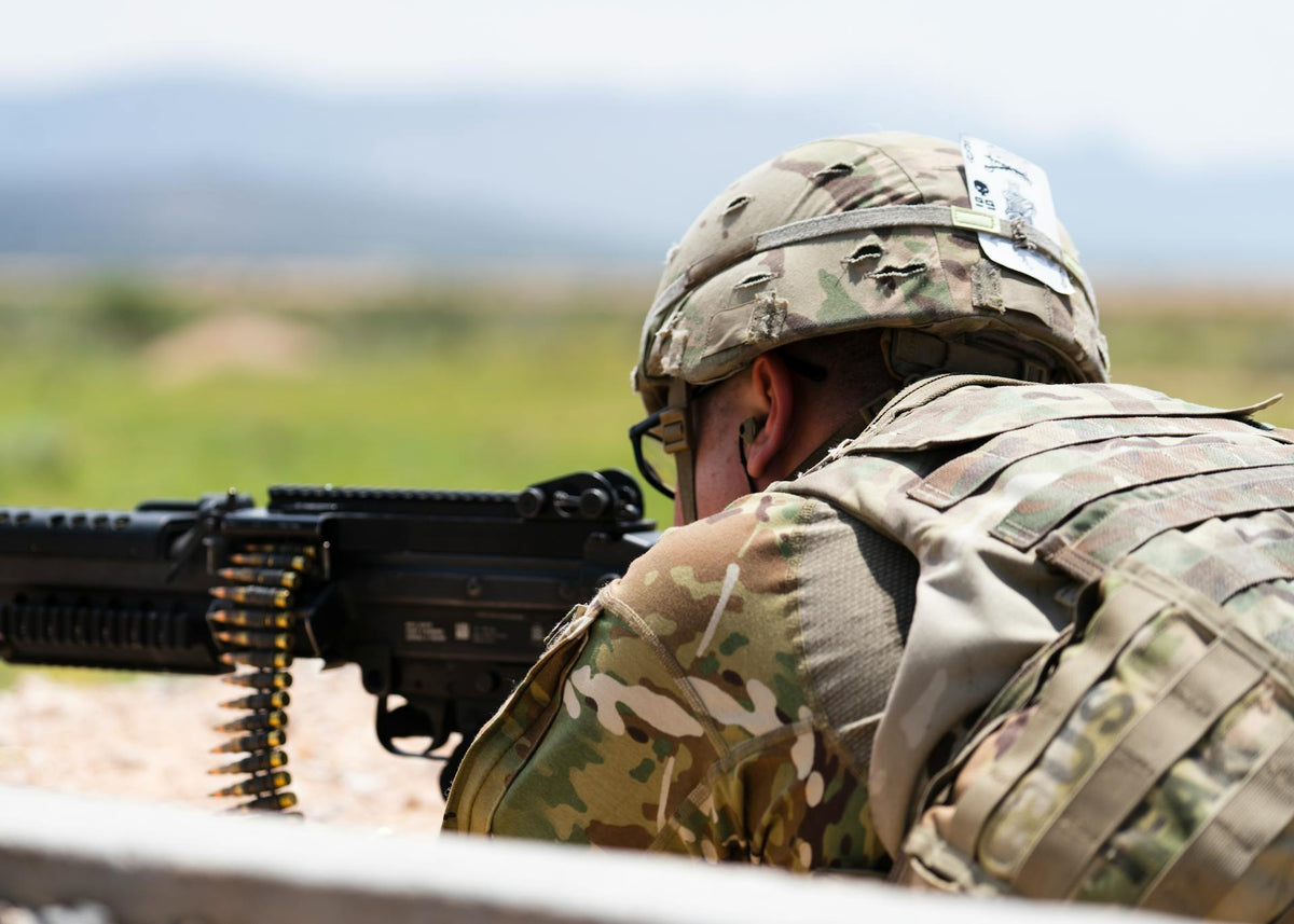 U.S. Army soldier in camouflage uniform aiming a machine gun while lying prone during field training, with ammunition belt visible and a blurred natural landscape in the background.